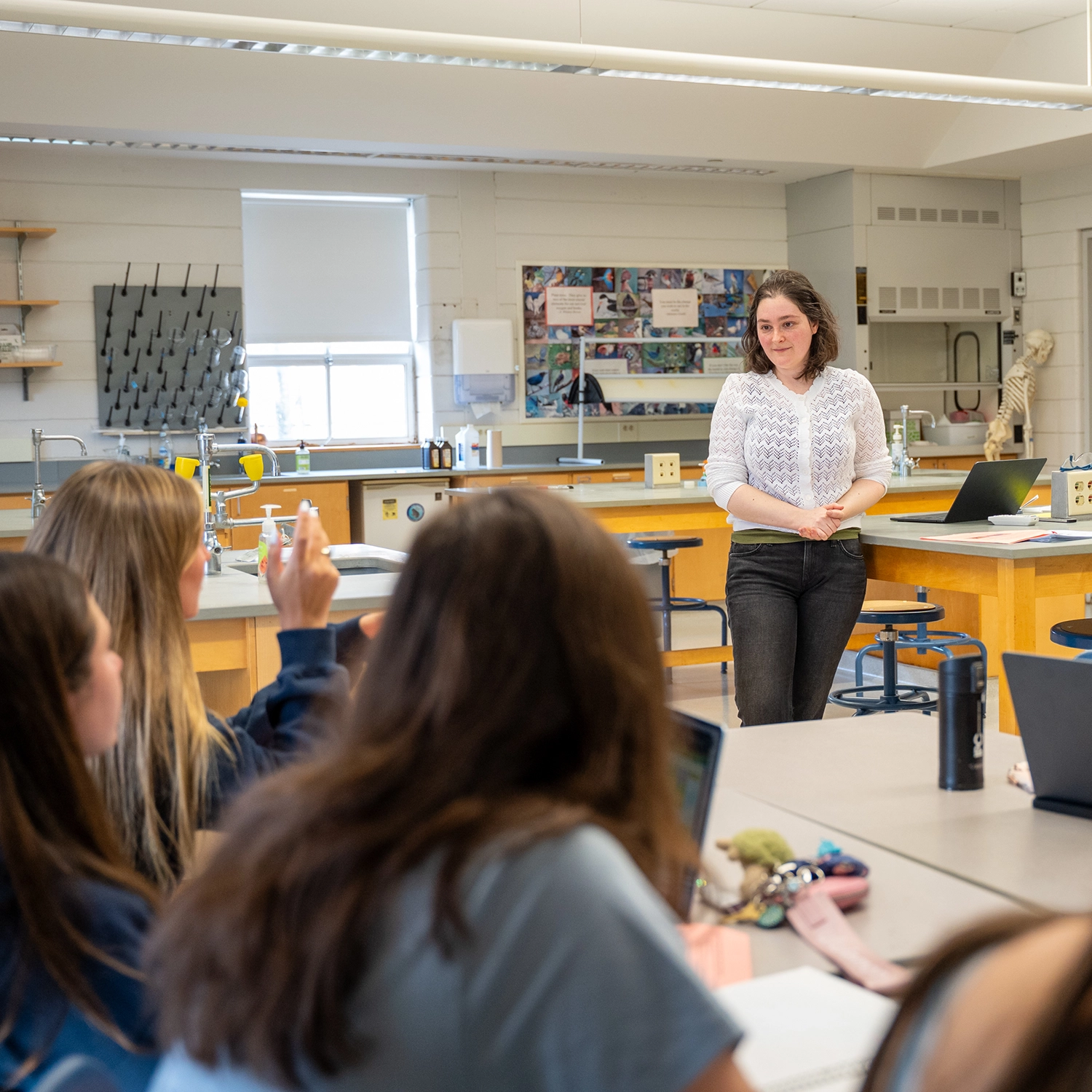A teacher stands at the front of a science classroom, engaging with students who are seated at tables; one student has their hand raised to ask a question.