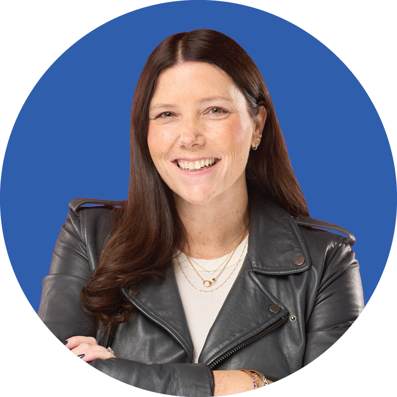 A woman with long brown hair, wearing a gray leather jacket and layered necklaces, smiles with arms crossed in front of a blue circular background.