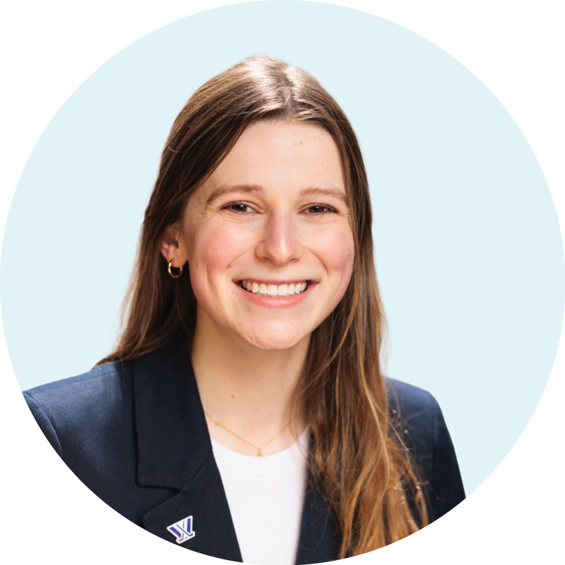 Smiling woman with long light brown hair wears a navy blazer with a pin and a white shirt, posing against a light blue background.