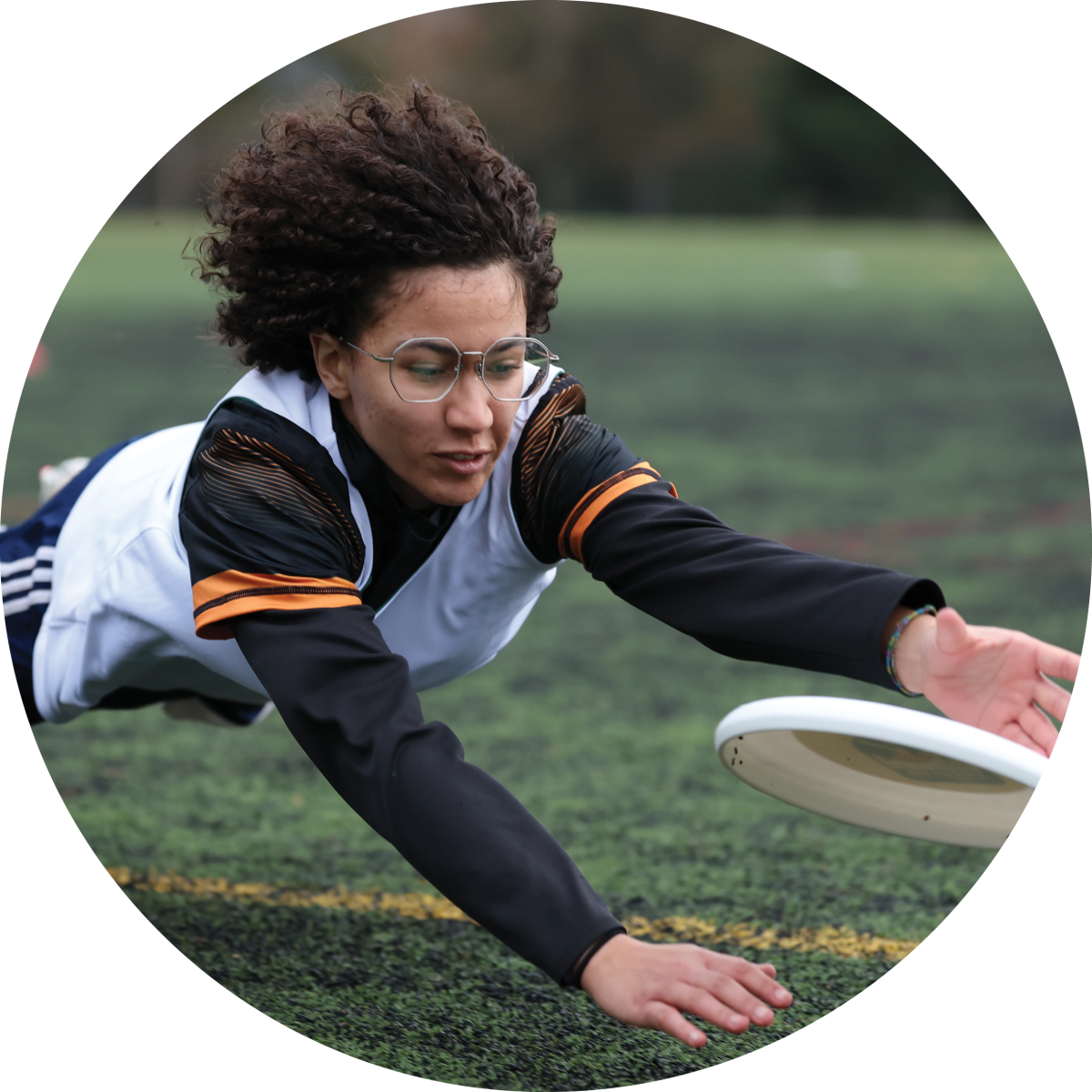 A person with curly hair and glasses dives to catch a white frisbee on a grassy field, wearing a sports jersey and black long-sleeve shirt.