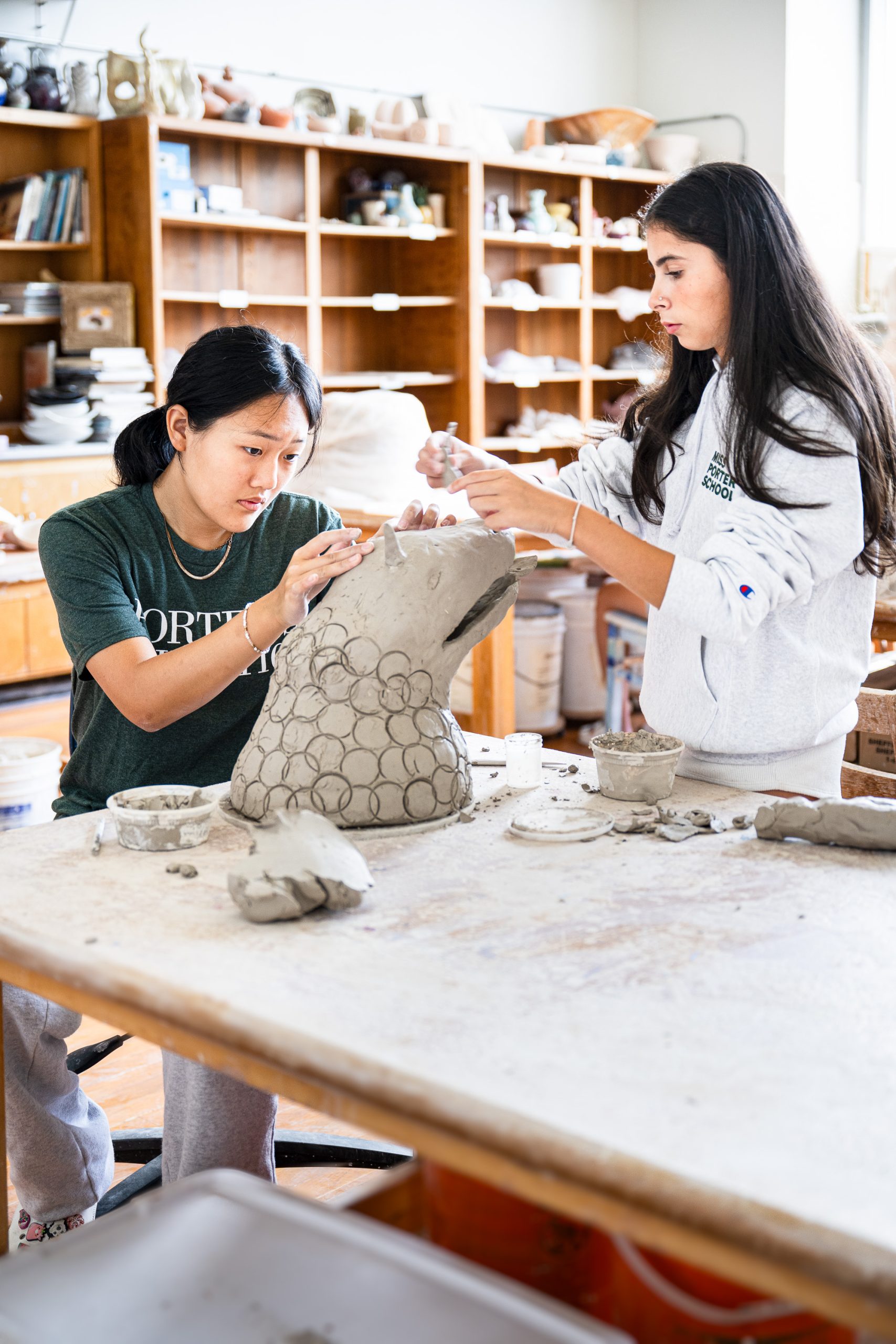 Two students sculpt a large clay project in an art classroom, focusing on adding circular textures to the piece. Shelves with pottery supplies and finished works are visible in the background.
