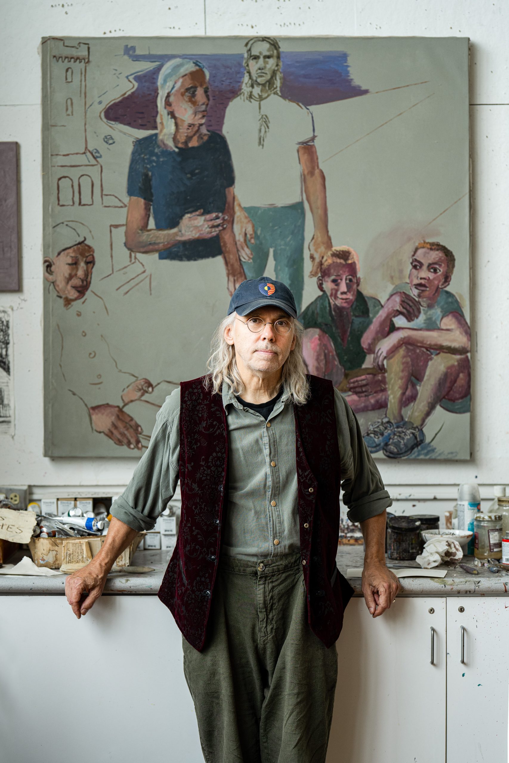 An older man with long gray hair, glasses, and a cap stands in an art studio, leaning against a table. Behind him is a large painting depicting four figures with expressive faces and postures.