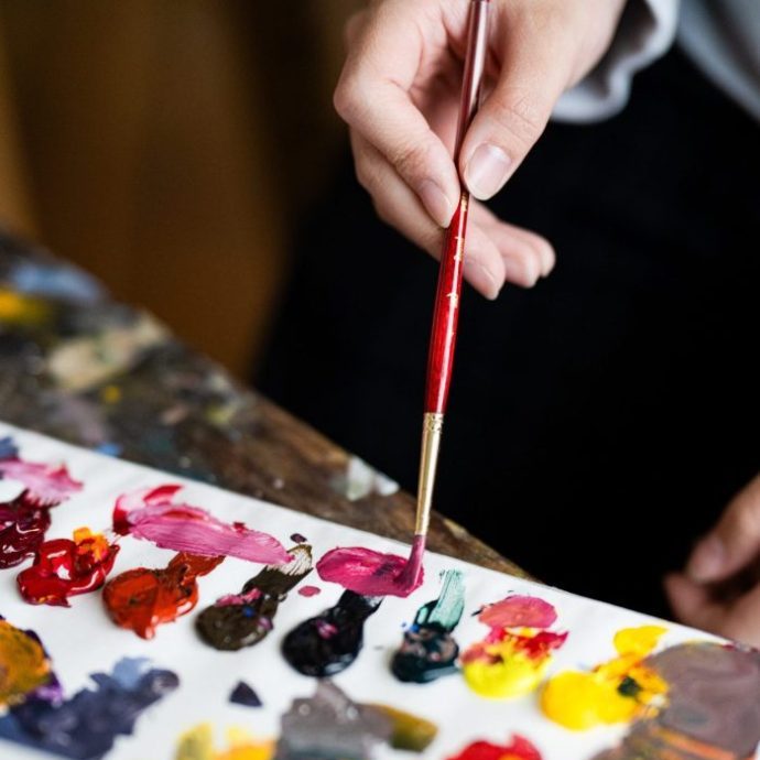 A hand holds a paintbrush, mixing colors on a palette covered with various bright paints.