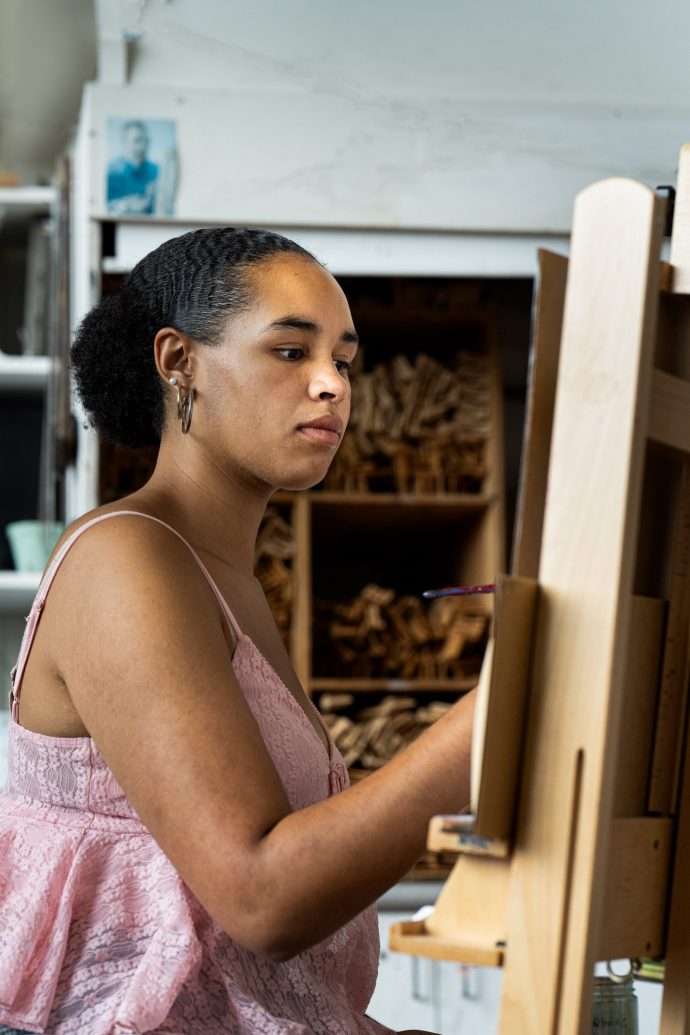 A woman in a pink lace top draws or paints on an easel in an art studio, looking focused. Shelves with art supplies and a small photo are in the background.