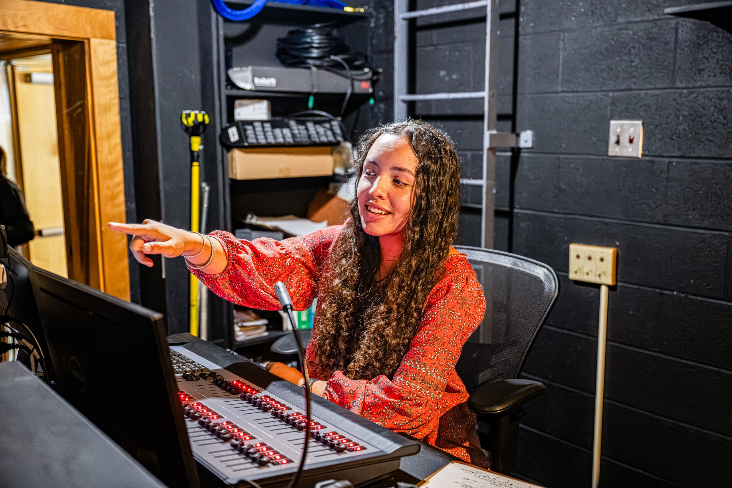 A young woman with long curly hair sits at a control panel, smiling and pointing at a screen in a backstage tech room with dark walls and equipment.