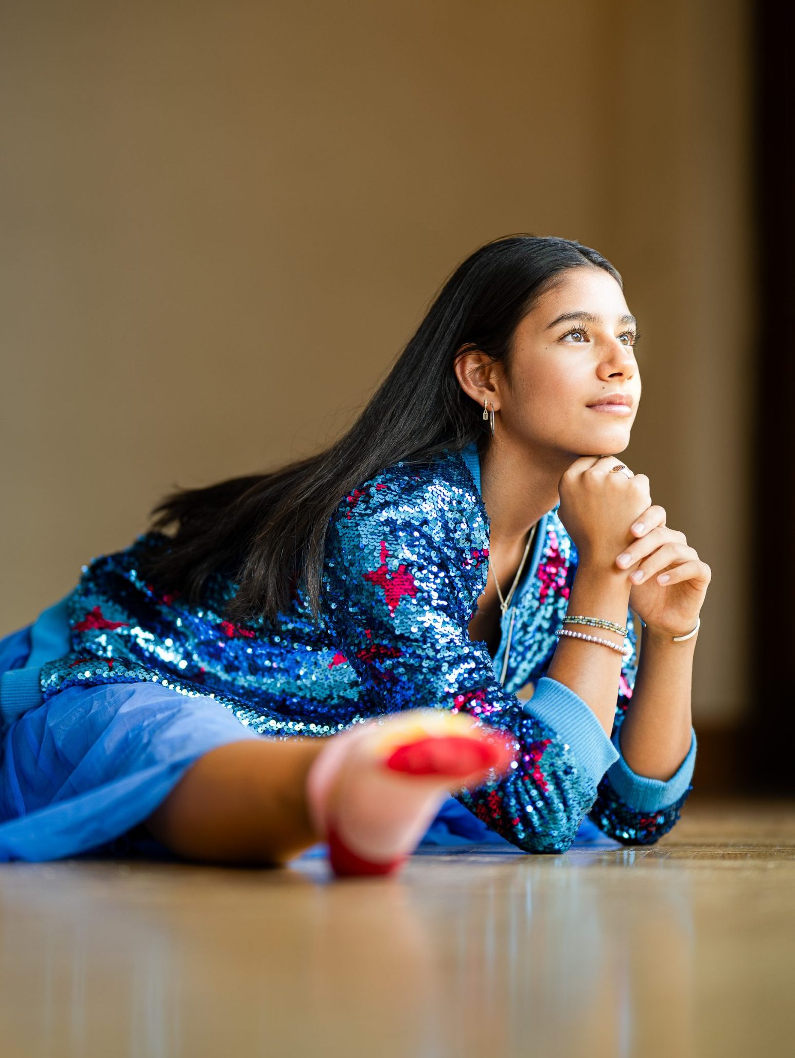 A woman in a blue sequin jacket and tulle skirt lies on the floor, resting on one arm and looking thoughtfully into the distance. Her hair is long and straight, and one leg is stretched out in front.