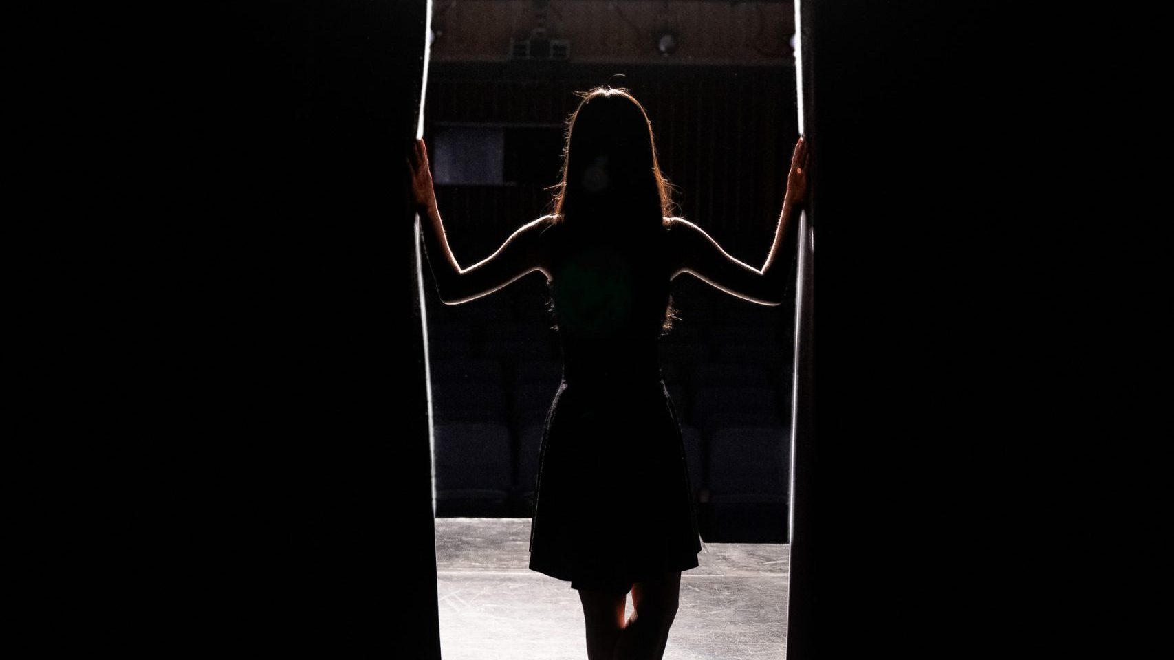 A person stands on stage, silhouetted by a spotlight, holding open stage curtains and looking out toward the audience.